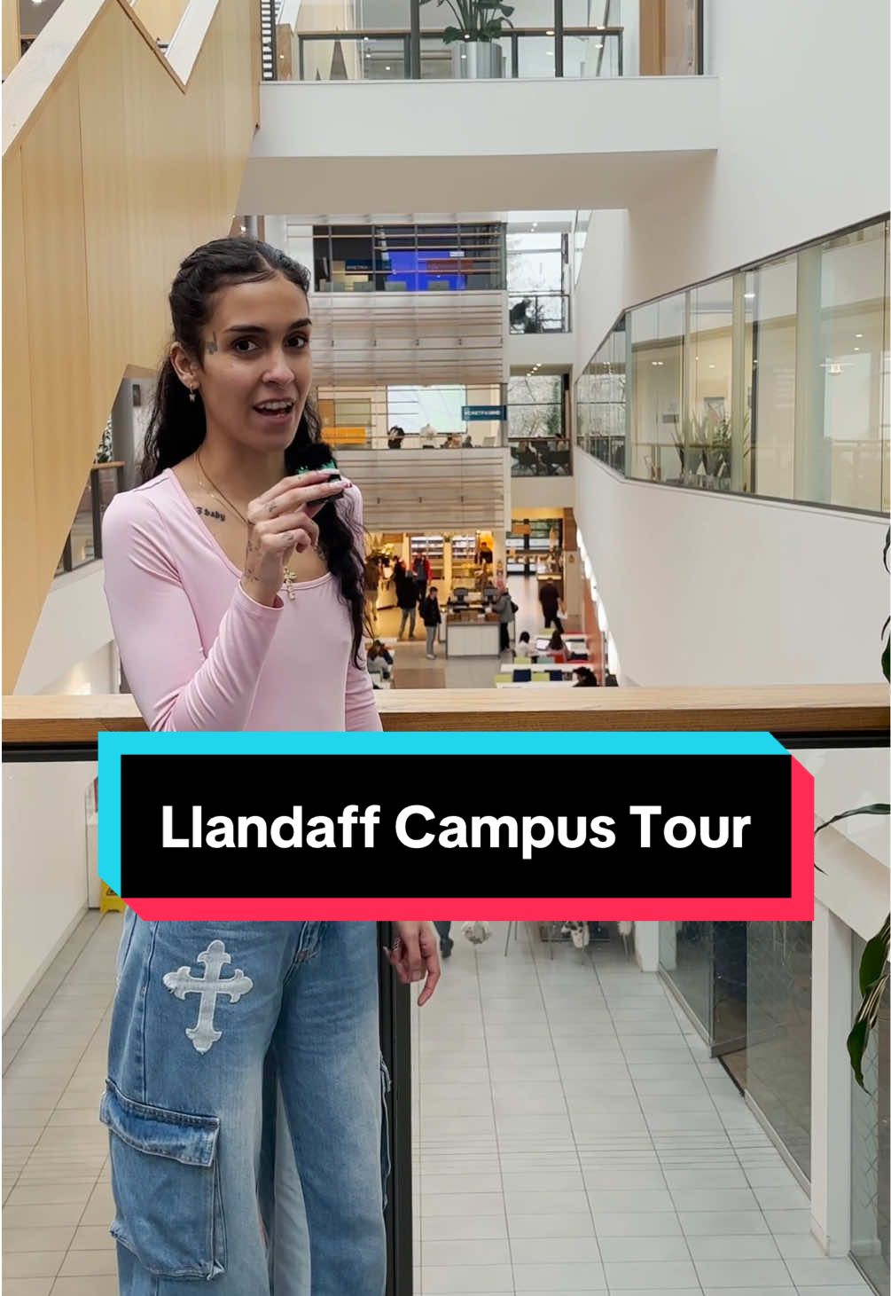 A young woman standing on a mezzanine in the Cardiff School of Management talks to camera, with the digital caption 