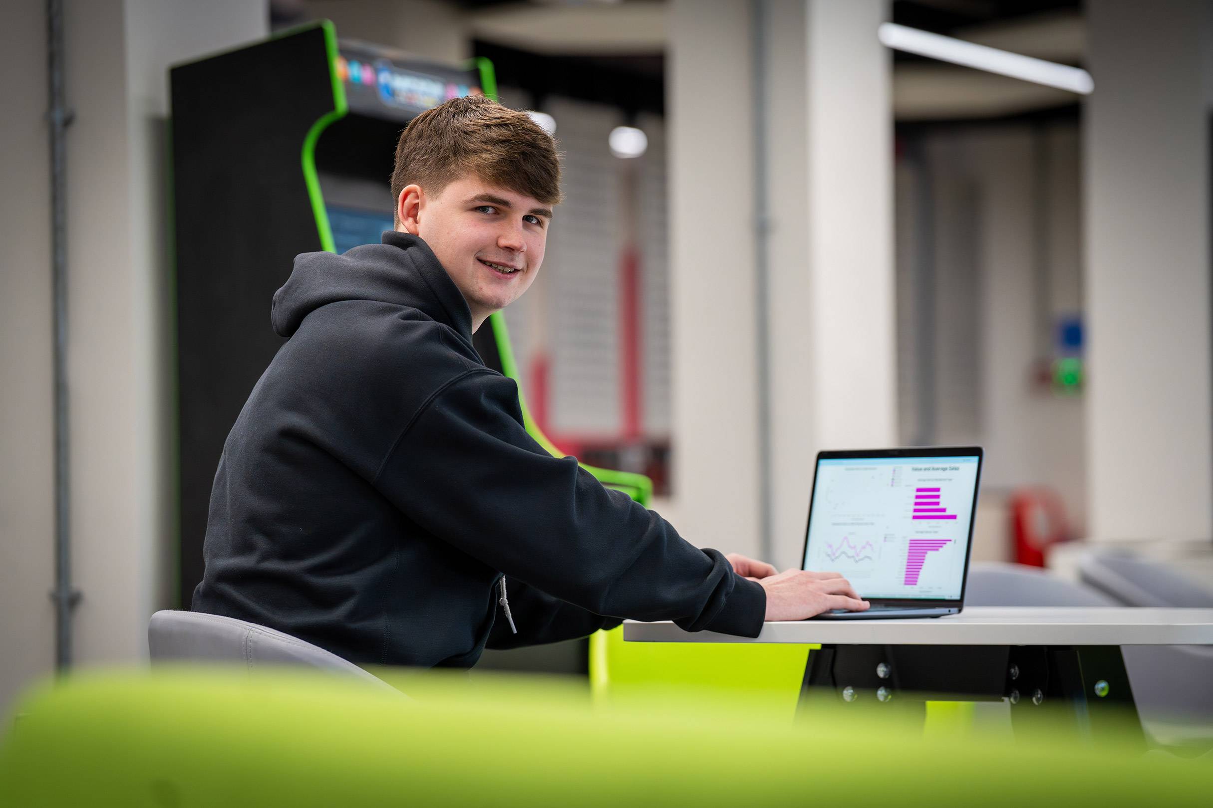 A young man works on a laptop at a table in an open workspace.