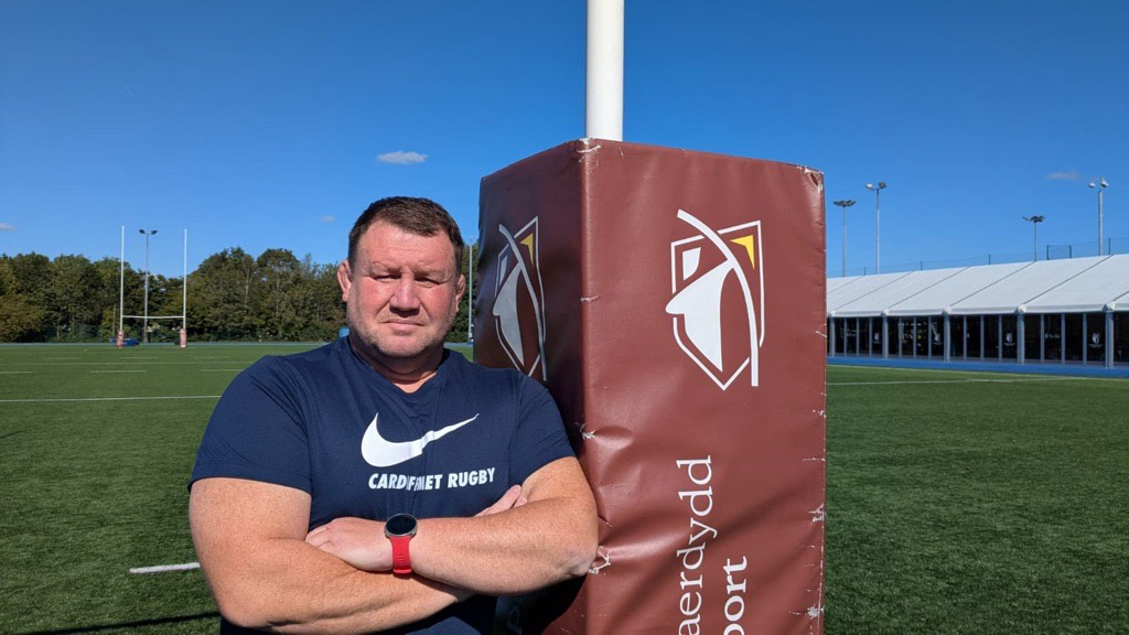 A man stands next to a rugby post, dressed in athletic clothing, with a rugby field visible in the background.